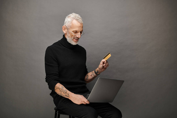 joyeux attrayant homme mature avec des lunettes à col roulé tenant carte de crédit et regardant ordinateur portable - Photo, image