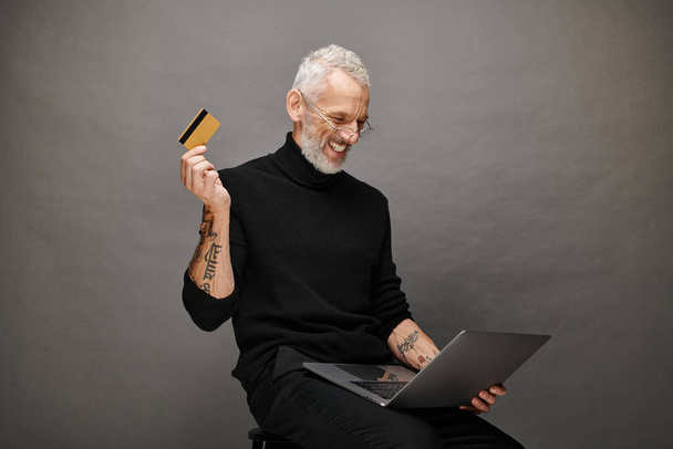 Boğazlı kazaklı, elinde kredi kartı olan ve dizüstü bilgisayarına bakan neşeli, çekici ve olgun bir adam. - Fotoğraf, Görsel