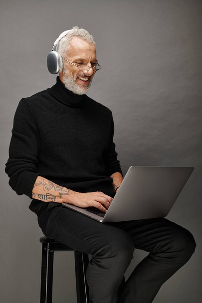 radosny przystojny mężczyzna w okularach i słuchawkach siedzący na krześle z laptopem i uśmiechnięty szczerze - Zdjęcie, obraz