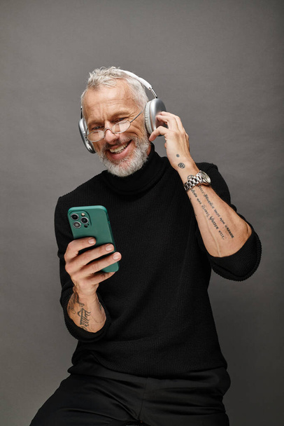 radosny atrakcyjny Dojrzałe mężczyzna model z broda i słuchawki w czarny golf patrząc na telefon - Zdjęcie, obraz