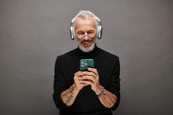 modelo masculino maduro atractivo alegre con barba y auriculares en cuello alto negro mirando el teléfono - Foto, imagen