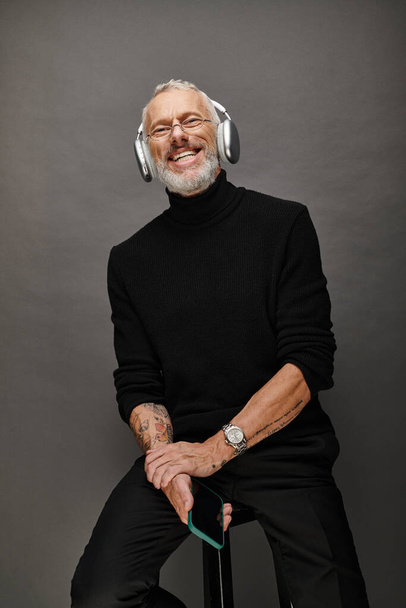 hombre maduro alegre en cuello alto negro con auriculares sosteniendo teléfono inteligente y mirando a la cámara - Foto, imagen