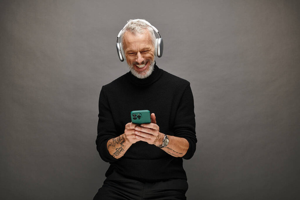 joyeux attrayant modèle masculin mature avec barbe et écouteurs en col roulé noir regardant le téléphone - Photo, image