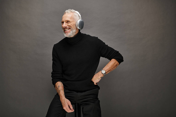joyful appealing mature man in voguish turtleneck with gray beard and headphones sitting on chair - Photo, Image