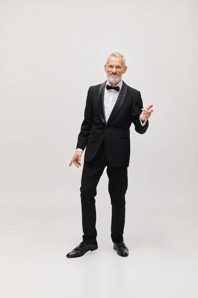 cheerful attractive gray bearded man with dapper stylish tuxedo dancing and smiling at camera - Foto, Bild
