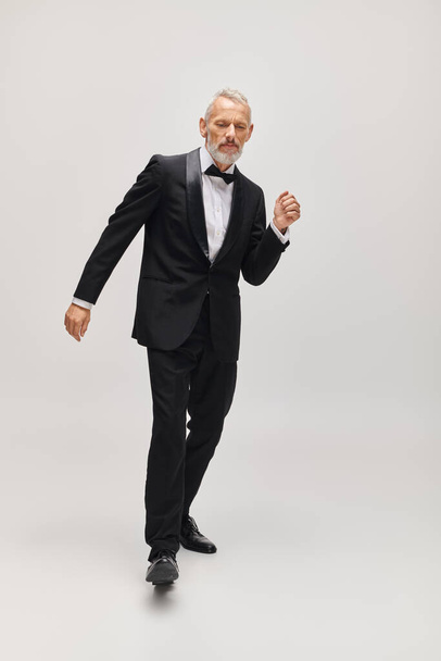 добре одягнений зрілий чоловік зі стильною краваткою-мішкою в voguish tuxedo танцює активно на сірому фоні - Фото, зображення