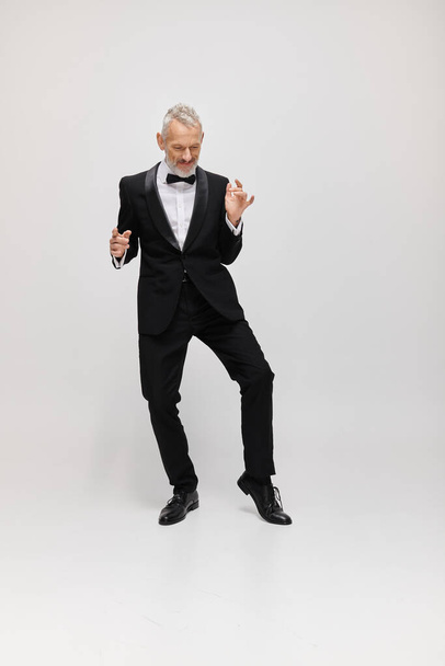 appealing merry mature male model with gray beard and bow tie in elegant tuxedo dancing actively - Photo, Image