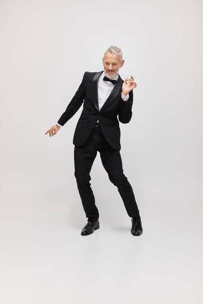 appealing merry mature male model with gray beard and bow tie in elegant tuxedo dancing actively - Photo, Image