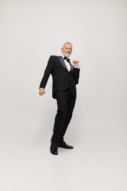 attractive happy mature man with gray beard and bow tie in elegant tuxedo dancing actively - Photo, Image
