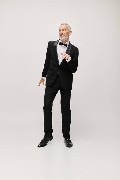 well dressed mature male model with bow tie in chic black tuxedo dancing actively on gray backdrop - Photo, Image