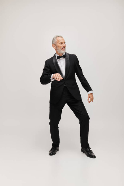 bem vestido modelo masculino maduro com laço gravata em elegante smoking preto dançando ativamente no fundo cinza - Foto, Imagem