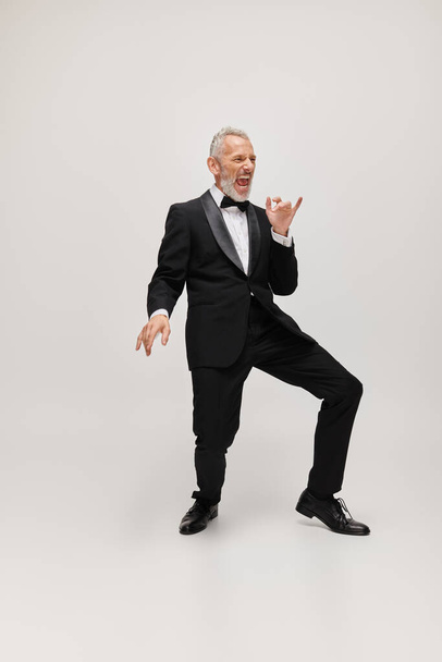 handsome joyful mature man with elegant dapper style with beard dancing happily on gray backdrop - Photo, Image