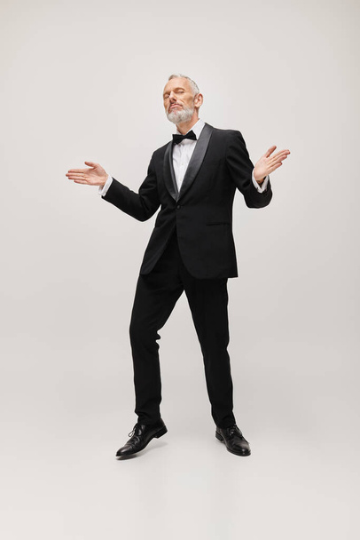 гарний вигляд зрілої чоловічої моделі з краваткою і бородою в елегантному тукседо танцює на сірому фоні - Фото, зображення