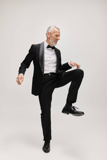 handsome joyful mature man with elegant dapper style with beard dancing happily on gray backdrop - Photo, Image