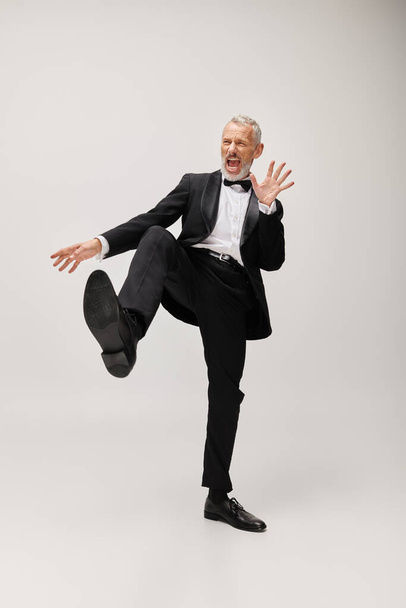 ansprechende lebenslustige reife Mann mit elegantem Stil mit Bart glücklich tanzen auf grauem Hintergrund - Foto, Bild