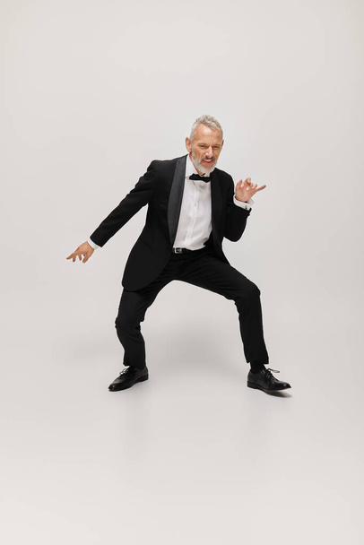 atraktivní veselý dospělý muž s elegantním elegantním elegantní styl s vousy tančí šťastně na šedém pozadí - Fotografie, Obrázek