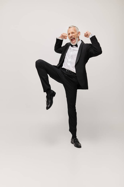 attractive jolly mature man with elegant dapper style with beard dancing happily on gray backdrop - Photo, Image