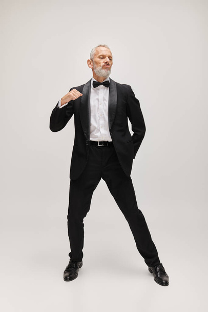 Çekici sakallı olgun adam güler yüzlü siyah smokinli papyonlu dans ederken hareket halinde poz veren - Fotoğraf, Görsel