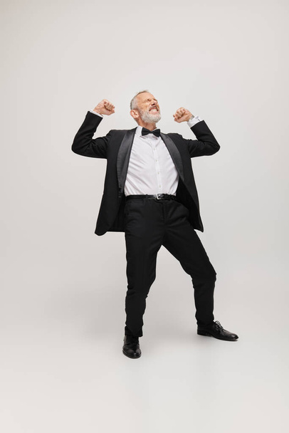 joyous good looking mature man in elegant suit with bow tie dancing happily on gray backdrop - Photo, Image