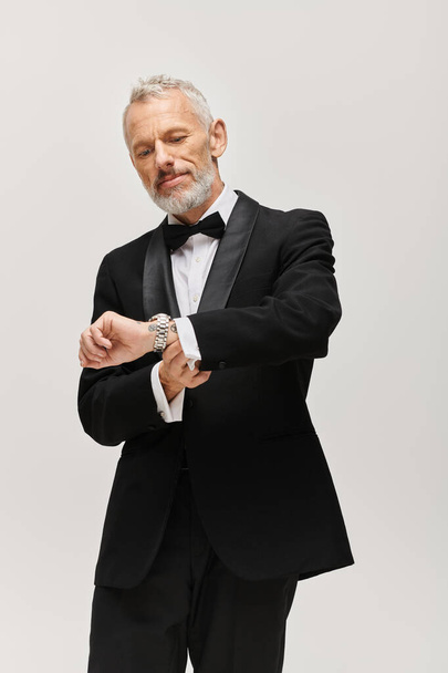 good looking mature bearded man with bow tie in elegant tuxedo posing and looking at wristwatch - Zdjęcie, obraz