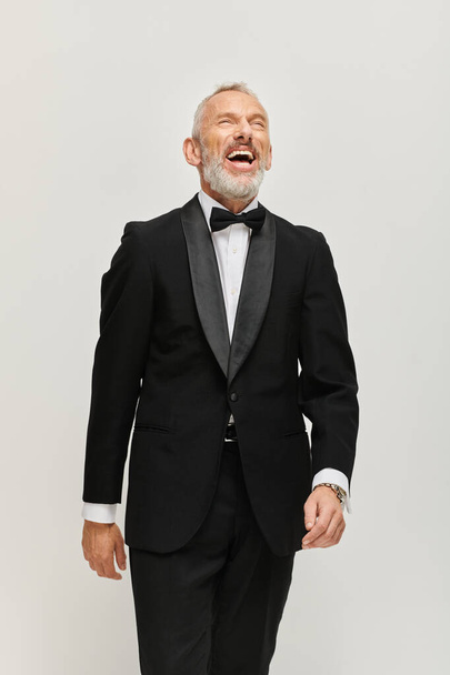 joyful good looking mature man with bow tie and gray beard in chic tuxedo smiling happily - Fotó, kép