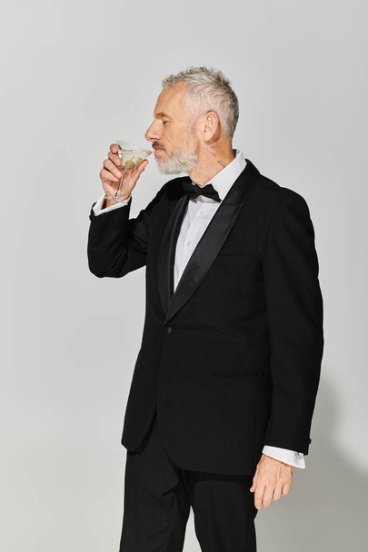hombre maduro de buen aspecto en elegante esmoquin con barba gris y pajarita bebiendo delicioso martini - Foto, imagen