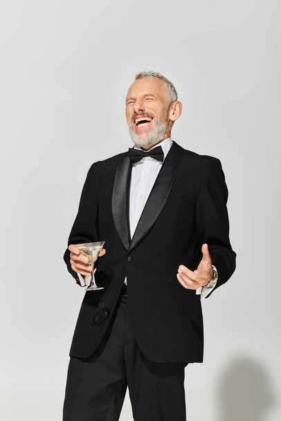 happy elegant mature man with beard in debonair tuxedo holding martini and smiling cheerfully - Foto, afbeelding