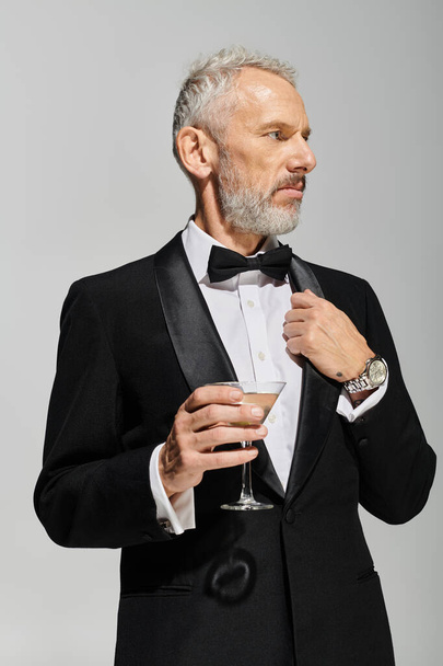 knappe bebaarde volwassen man in elegante smoking met vlinderdas met martini en wegkijkend - Foto, afbeelding