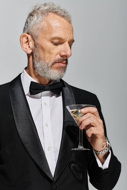 vonzó szakállas érett férfi elegáns szmokingban csokornyakkendővel tartó pohár martini olívabogyóval - Fotó, kép