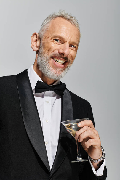 alegre buen aspecto hombre maduro en elegante esmoquin sosteniendo vaso de martini y sonriendo a la cámara - Foto, Imagen