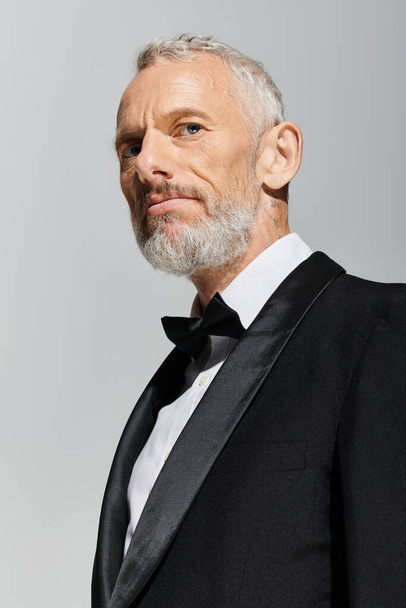 good looking elegant mature man with beard wearing fashionable tuxedo and looking at camera - Foto, afbeelding