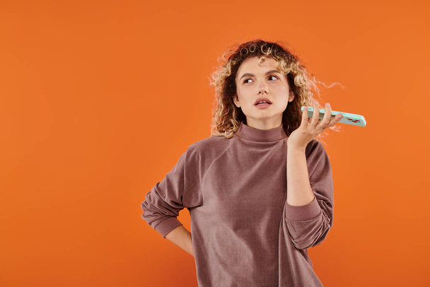mujer rizada reflexiva en color moca mensaje de voz de grabación de cuello alto en el teléfono inteligente en naranja - Foto, Imagen