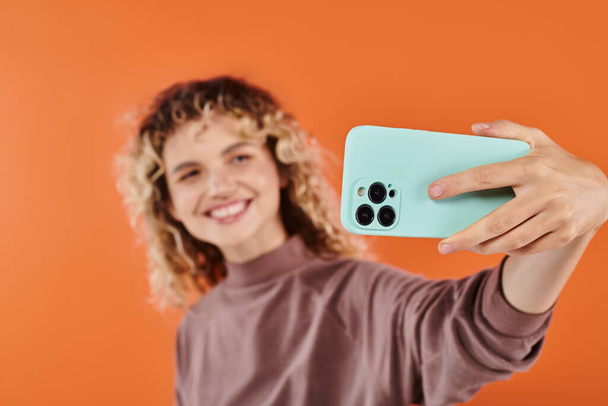 lächelnde trendige Frau mit welligem Haar macht Selfie auf Handy auf verschwommenem orangefarbenem Hintergrund - Foto, Bild