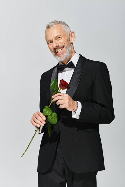 カメラで赤いバラと微笑んでいるエレガントなタキシードのひげを持つ魅力的な陽気な成熟した男 - 写真・画像