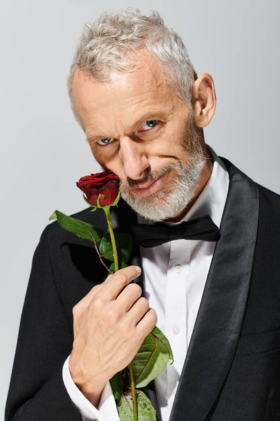 appealing fashionable mature man with beard in tuxedo holding red rose and looking at camera - Photo, Image