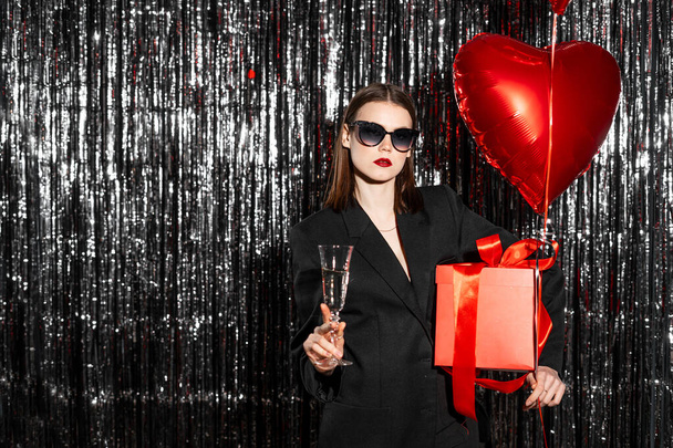A beautiful young woman in black suit with heart shape air balloon and gift on a tinsel background. Woman on Valentine's Day. Symbol of love. Copy space - Photo, Image