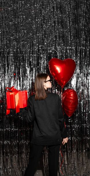 Стильная молодая женщина с воздушным шаром в форме сердца на серебристом ярком фоне, вид сзади. День святого Валентина - Фото, изображение