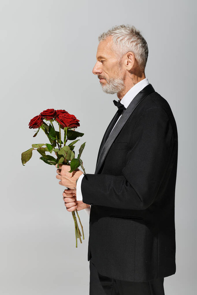 elegant good looking mature man in voguish black tuxedo holding red roses bouquet in hands - Photo, Image