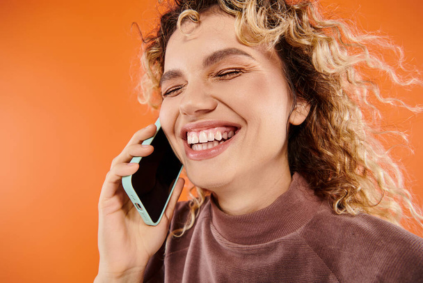 überglücklich lockige Frau lacht während Handy-Gespräch auf leuchtend orangefarbenem Hintergrund, Glück - Foto, Bild