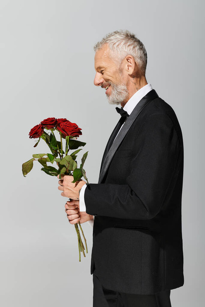 Веселый, красивый, зрелый мужчина в модном черном смокинге с букетом красных роз в руках - Фото, изображение