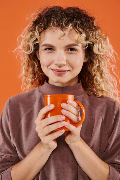 mujer sonriente con el pelo rizado y taza de café de la mañana mirando a la cámara en naranja, rutina diaria - Foto, Imagen