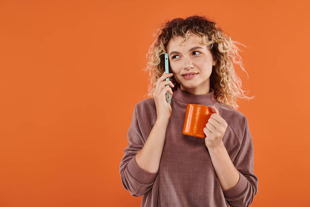 boldog göndör nő csésze reggeli kávé beszél okostelefon világos narancs háttér - Fotó, kép