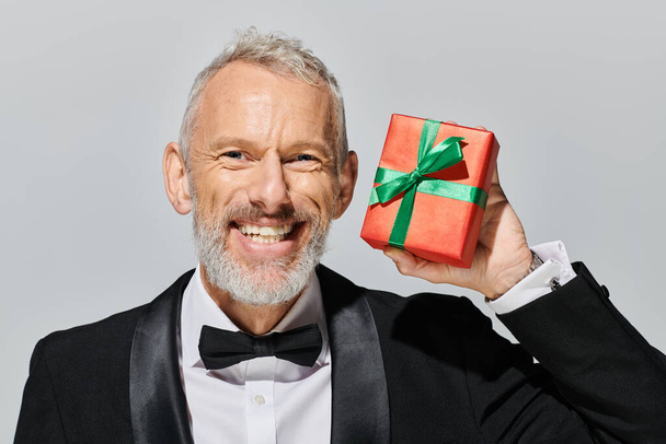 весёлый, симпатичный, зрелый мужчина с бородой в смокинге держит подарок и улыбается в камеру. - Фото, изображение