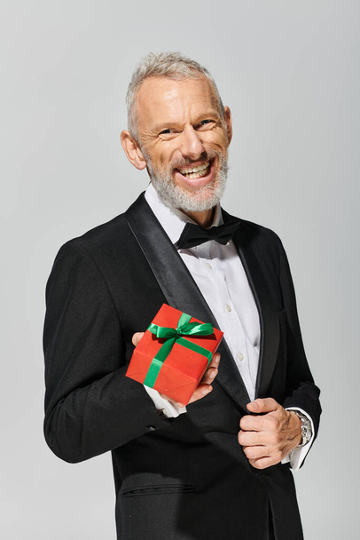 freudig gut aussehender Debonair reifer Mann mit Bart im Smoking hält Geschenk in der Hand und lächelt in die Kamera - Foto, Bild