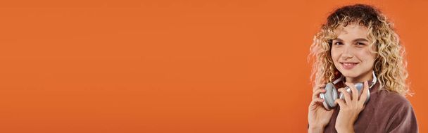 heureuse femme bouclée à col roulé élégant avec écouteurs sans fil regardant la caméra sur orange, bannière - Photo, image
