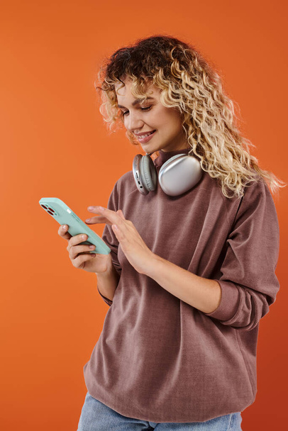 Kablosuz kulaklıklı neşeli kıvırcık kadın turuncu arka planda cep telefonuyla mesajlaşıyor. - Fotoğraf, Görsel