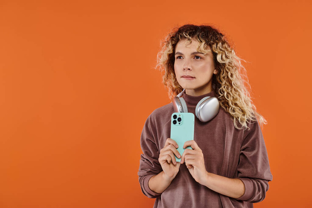 στοχαστική σγουρή γυναίκα με ασύρματα ακουστικά και κινητό τηλέφωνο κοιτάζοντας μακριά σε πορτοκαλί φόντο - Φωτογραφία, εικόνα