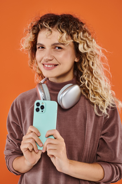 radostná kudrnatá žena s bezdrátovými sluchátky a mobilním telefonem při pohledu do kamery na oranžovém pozadí - Fotografie, Obrázek