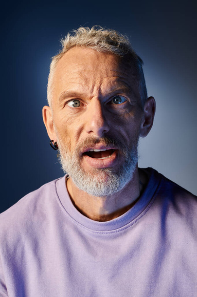 good looking mature man with beard and casual fashionable attire grimacing actively on blue backdrop - Photo, Image