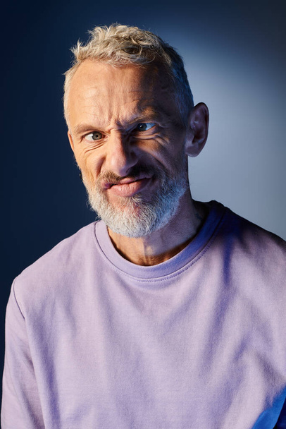 good looking mature man with beard and casual fashionable attire grimacing actively on blue backdrop - Photo, Image